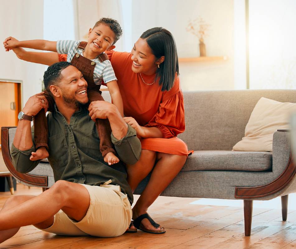 Image of a woman sitting on a couch, with a man sitting on the floor in front of her, holding a child on his shoulders